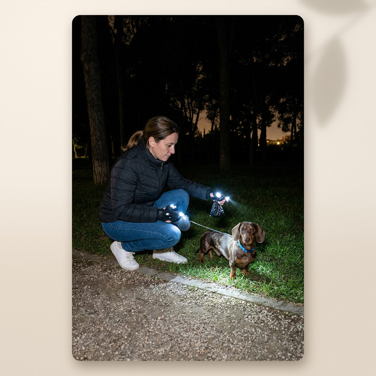 Mujer recogiendo lo de su teckel de noche en el parque con la luz de los guantes LED AMIKART